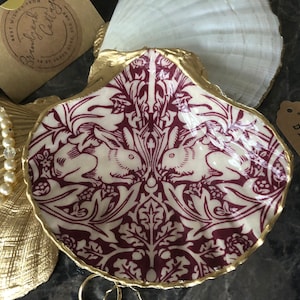May include: A white and burgundy trinket dish shaped like a seashell with a gold rim. The dish features a repeating pattern of stylised rabbits and leaves. The dish is labelled "trinket dish".