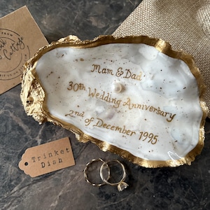Könnte beinhalten: Eine Austernschalen-Schmuckschale mit weißem Inneren und goldfarbenen Rändern. Im Inneren befindet sich eine goldene Kalligrafie mit der Aufschrift "Mam & Dad 30th Wedding Anniversary 2nd of December 1995" sowie eine kleine Perle. Zwei Goldringe liegen daneben.