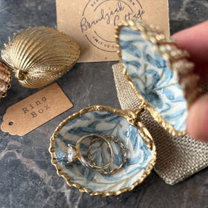 May include: A seashell ring box with a blue and white floral pattern, holding two rings. The box is open, revealing the rings inside. A gold-colored seashell and a tag that says "Ring Box" are also visible.