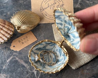 May include: A seashell ring box with a blue and white floral pattern, holding two rings. The box is open, revealing the rings inside. A gold-colored seashell and a tag that says "Ring Box" are also visible.