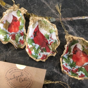 May include: Three gold-leafed oyster shell ornaments featuring a cardinal bird and holly design. The ornaments are decorated in red, green and white.