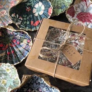 May include: A set of five decorative ring dishes made from seashells, each with a different floral pattern. The dishes are packaged in a brown box with a label that reads "Ring Dish".