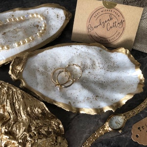 May include: Handmade oyster shell trinket trays with gold trim and speckled accents. One tray holds gold rings, another a pearl bracelet. A gold watch and a tag that says "Trinket Tray" are also visible.