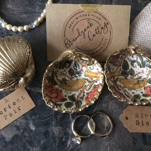 Könnte beinhalten: Zwei goldfarbene Keramik-Ringteller mit einem floralen und Vogel-Design. Die Teller sind offen und liegen auf einer dunkelgrauen Oberfläche. In den Tellern befinden sich zwei goldene Ringe und ein Diamantring. Ein kleines braunes Etikett mit dem Text "Ring Dish" befindet sich auf der rechten Seite des Bildes. Ein kleines braunes Etikett mit dem Text "A perfect pair" befindet sich auf der linken Seite des Bildes.