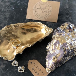 May include: Two gold oyster shell trinket dishes. One dish is filled with gold leaf and the other is a natural oyster shell with gold leaf accents. The dishes are on a dark grey surface with a small tag that says "Trinket Dish".