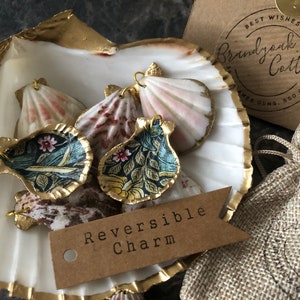 May include: A set of reversible shell charms with a floral pattern. The charms are displayed on a white seashell with gold accents. The text "Reversible Charm" is written on a brown tag.