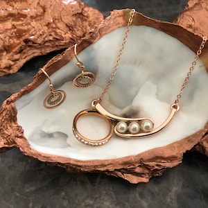 Könnte beinhalten: Ein roségoldenes Schmuckset mit einer Halskette mit drei kleinen Perlen, passenden Ohrringen und einem Ring mit klaren Kristallen. Der Schmuck wird auf einer weiß-kupferfarbenen dekorativen Austernschale präsentiert.