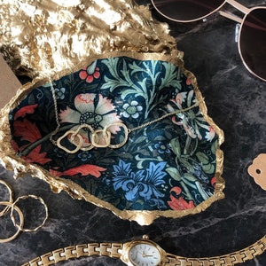 Pu&ograve; includere: Un vassoio decorativo per bigiotteria con un motivo floreale nei toni del blu, verde e rosa, bordato d'oro. Una collana, anelli e un orologio dorati sono esposti nel piatto. Sono visibili anche occhiali da sole e un'etichetta con la scritta "Trinket Dish".