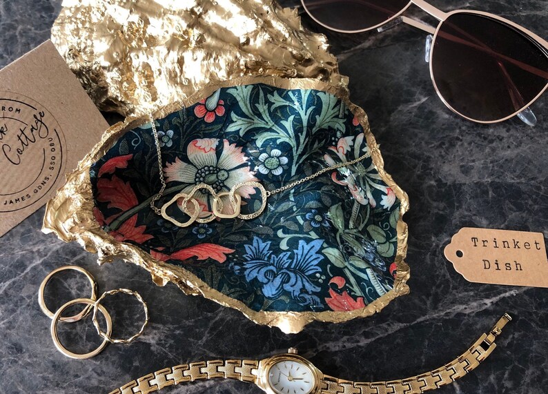 Pu&ograve; includere: Un vassoio decorativo per bigiotteria con un motivo floreale nei toni del blu, verde e rosa, bordato d'oro. Una collana, anelli e un orologio dorati sono esposti nel piatto. Sono visibili anche occhiali da sole e un'etichetta con la scritta "Trinket Dish".