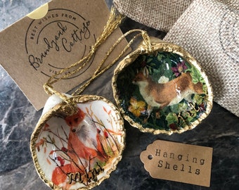 May include: Two gold-trimmed seashells with a fox design. The shells are hanging from gold cords with tassels. The shells are on a dark surface with a brown tag that says "Hanging Shells".