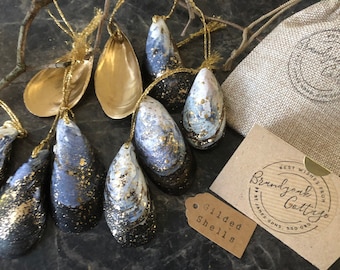 May include: A set of eight black and white painted seashells with gold accents. The shells are strung with gold twine and have a tag that reads "Gilded Shells".