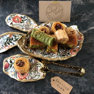 May include: A white ceramic serving dish shaped like a rabbit with gold trim and a floral pattern. The dish is filled with a variety of Middle Eastern sweets, including baklava and pistachio-filled pastries. A pair of gold serving tongs and a brown tag with the text "Serving Dish" are also visible.