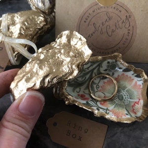 May include: A gold oyster shell ring box with a floral design and a gold ring inside. The box is sitting on a brown surface with a tag that says "A perfect pair".