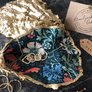 Pu&ograve; includere: Un portagioie a forma di conchiglia di ostrica dorata con interno a fantasia floreale. Una collana d'argento con anelli intrecciati &egrave; all'interno. Un'etichetta recita "Trinket Dish". Una carta recita "Best Wishes From Brandjack Cottage".