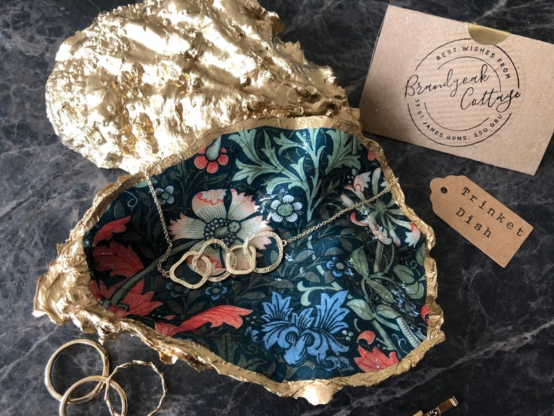 Pu&ograve; includere: Un portagioie a forma di conchiglia di ostrica dorata con interno a fantasia floreale. Una collana d'argento con anelli intrecciati &egrave; all'interno. Un'etichetta recita "Trinket Dish". Una carta recita "Best Wishes From Brandjack Cottage".