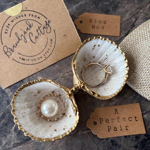Puede incluir: Un joyero en forma de concha con borde dorado, que contiene dos anillos. Uno tiene una piedra transparente, el otro una perla. Las etiquetas indican "Ring Box" y "A Perfect Pair". Tambi&eacute;n hay una tarjeta con texto.