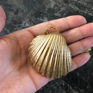 May include: A gold-colored seashell pendant with a textured surface and a woven gold-colored loop for hanging. The pendant is held in a hand against a dark background.