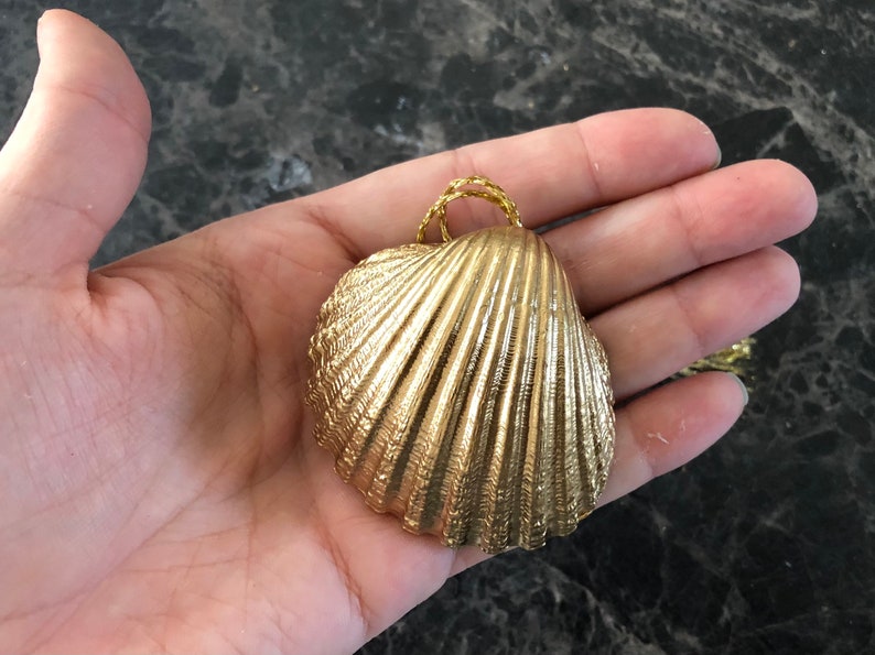 May include: A gold-colored seashell pendant with a textured surface and a woven gold-colored loop for hanging. The pendant is held in a hand against a dark background.