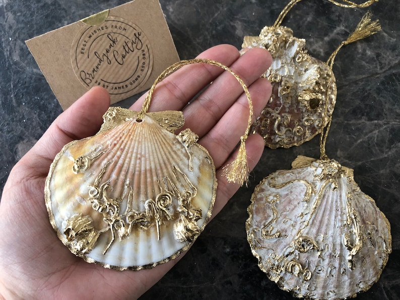 May include: Three gold-painted seashell ornaments with a gold tassel hanging from each. The seashells are decorated with gold paint and have a natural white and brown color.
