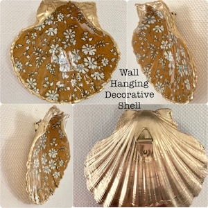 May include: A gold-painted seashell with a floral pattern on the inside. The shell has a hanging loop on the back and is a decorative wall hanging.