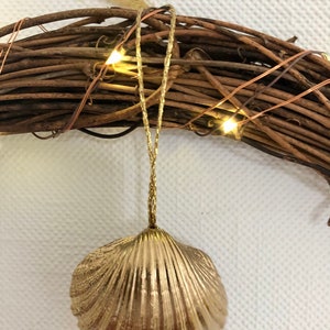 May include: A gold-colored seashell ornament hangs from a grapevine wreath. The seashell has a textured surface and is suspended by a gold cord. The wreath is adorned with small, warm white lights.