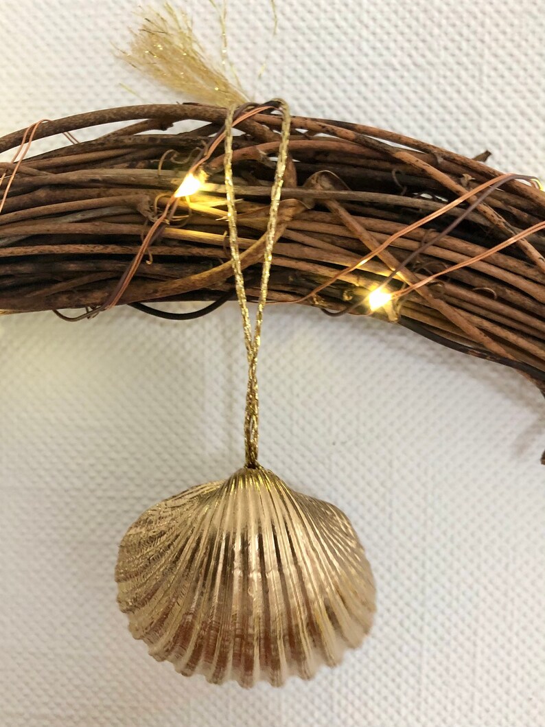 May include: A gold-colored seashell ornament hangs from a grapevine wreath. The seashell has a textured surface and is suspended by a gold cord. The wreath is adorned with small, warm white lights.