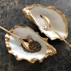 Op de afbeelding: Twee witte oesterschelp zout- en peperstrooiers met gouden accenten. Elke strooier heeft een bijpassende gouden lepel met een heldere kristallen handvat.