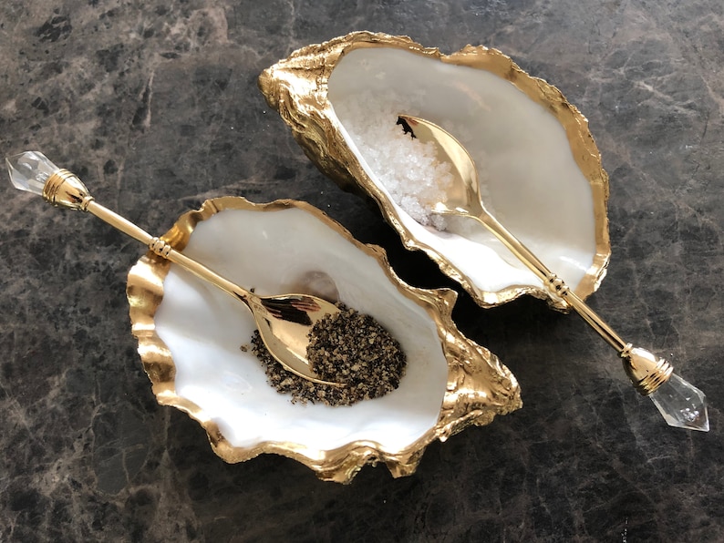 Op de afbeelding: Twee witte oesterschelp zout- en peperstrooiers met gouden accenten. Elke strooier heeft een bijpassende gouden lepel met een heldere kristallen handvat.