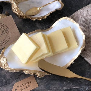 May include: A white oyster shell butter dish with gold accents, filled with butter pats and a gold butter knife. The dish is on a dark surface with a brown burlap napkin.