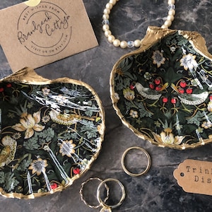 May include: Two gold-rimmed seashell trinket dishes with a black background and a floral pattern featuring birds and red berries. The dishes are on a grey surface with gold rings and a brown tag that says "Trinket Dish".