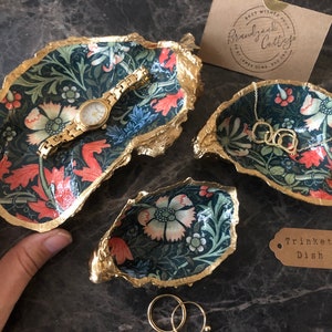 Pu&ograve; includere: Set di tre vassoi decorativi per bigiotteria con motivo floreale nei toni del verde, rosso e crema. I bordi sono rifiniti in oro. Un orologio e una collana d'oro sono esposti. Un'etichetta recita "Trinket Dish".