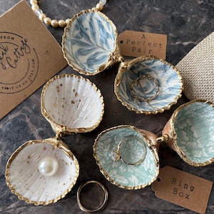 Puede incluir: Cajas de anillos decorativas con forma de concha marina, con bordes dorados e interiores estampados. Una caja contiene una perla, otras contienen anillos. Una etiqueta dice "A Perfect Pair". Una tarjeta de visita dice "Best Wishes From Brandyrk Cottage".