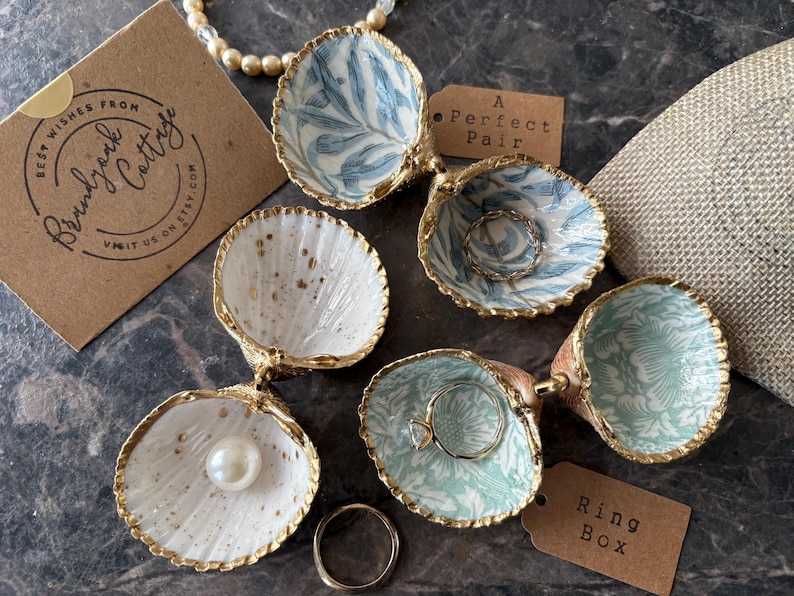 Puede incluir: Cajas de anillos decorativas con forma de concha marina, con bordes dorados e interiores estampados. Una caja contiene una perla, otras contienen anillos. Una etiqueta dice "A Perfect Pair". Una tarjeta de visita dice "Best Wishes From Brandyrk Cottage".