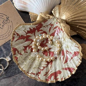 May include: A decorative seashell trinket dish with a red floral pattern and gold trim, holding a pearl bracelet. Two rings sit nearby. A gold-painted seashell and a tag that says "Trinket Dish" are also visible.