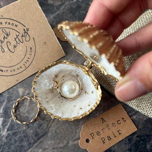 May include: A decorative seashell ring holder with a gold trim, containing a pearl ring. A gold twisted ring and a tag reading "A Perfect Pair" are also present. The brand name "Brandyork Cottage" is printed on a brown card.