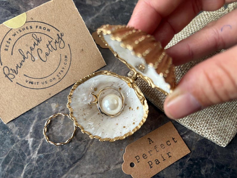 Puede incluir: Un joyero en forma de concha con borde dorado, que contiene un anillo de perlas. Tambi&eacute;n se ve un anillo dorado retorcido y una etiqueta que dice "A Perfect Pair". La marca "Brandyork Cottage" est&aacute; impresa en una tarjeta marr&oacute;n.