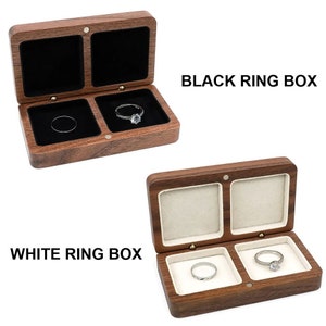 Puede incluir: Dos cajas de anillos de madera, una con forro de terciopelo negro y otra con forro de terciopelo blanco. Ambas cajas tienen dos compartimentos para anillos. El texto "BLACK RING BOX" está sobre la caja negra y "WHITE RING BOX" está sobre la caja blanca.