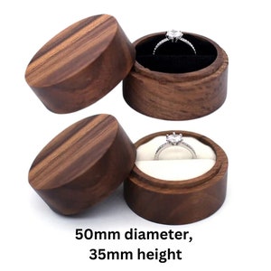 Peut inclure: Une boîte à bague ronde en bois avec un couvercle. La boîte mesure 50 mm de diamètre et 35 mm de hauteur. La boîte est en bois foncé avec un motif de grain naturel. Le couvercle est ouvert et une bague en argent avec une pierre transparente est à l'intérieur.
