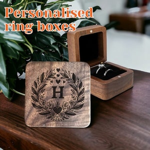 Puede incluir: Cajas de anillos de madera, una abierta con anillos dentro. Las cajas son marrones con un interior oscuro. Una caja tiene el texto "Personalised ring boxes". Un cuadrado de madera con la letra "H" está grabado con un diseño floral.