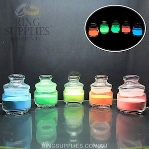 May include: Five small, clear glass jars with lids, each filled with a different colored powder: blue, green, yellow-green, orange, and pink. A smaller inset image shows the jars glowing in the dark.