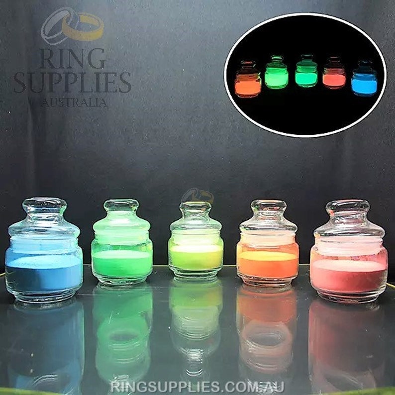 May include: Five small, clear glass jars with lids, each filled with a different colored powder: blue, green, yellow-green, orange, and pink. A smaller inset image shows the jars glowing in the dark.