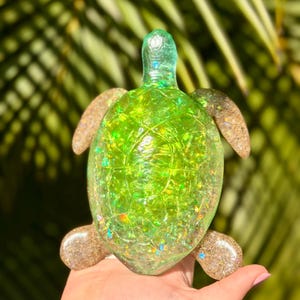 May include: A decorative turtle figurine with a translucent green body filled with glitter. The turtle's head and flippers are a sandy brown color. The figurine is held in a hand against a blurred green background.