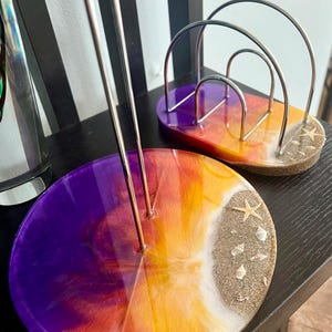 May include: Two round resin coasters with a sunset gradient design. The coasters feature a white beach with sand and seashells. One coaster has a metal wire stand for holding paper.