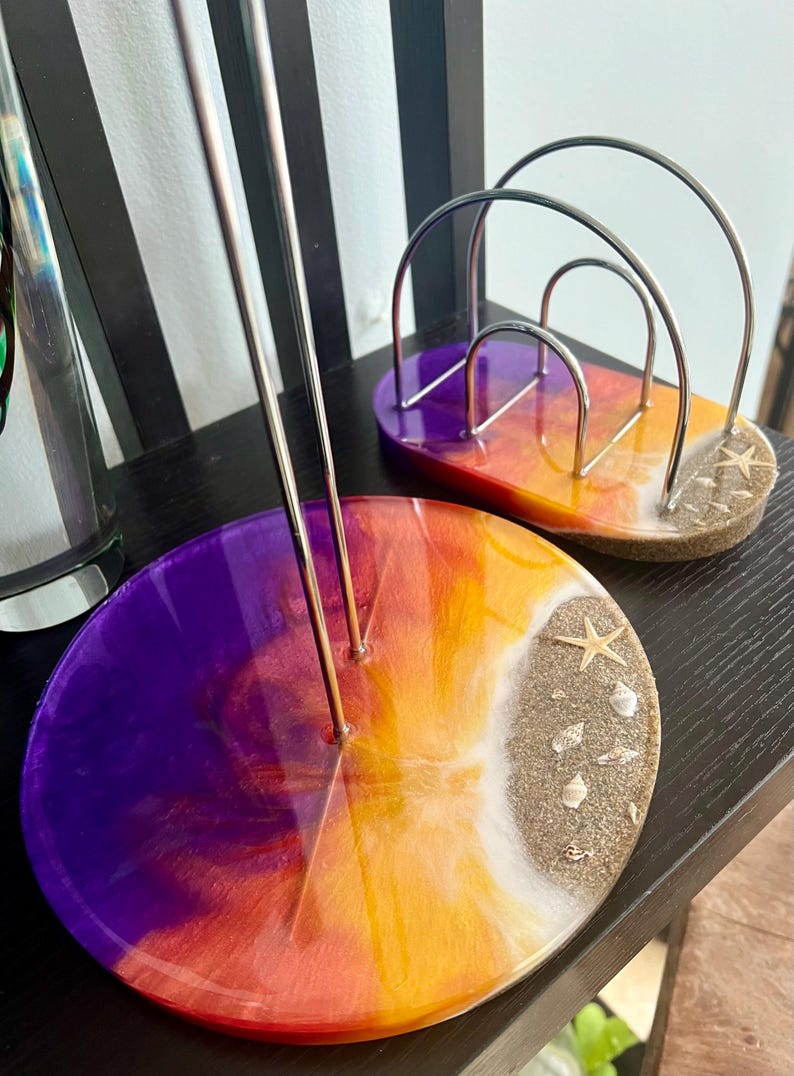 May include: Two round resin coasters with a sunset gradient design. The coasters feature a white beach with sand and seashells. One coaster has a metal wire stand for holding paper.