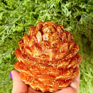 May include: A decorative pinecone-shaped object crafted from a glossy, translucent resin material. The surface features a vibrant copper-orange hue infused with numerous reflective gold and orange glitter particles that catch the light.