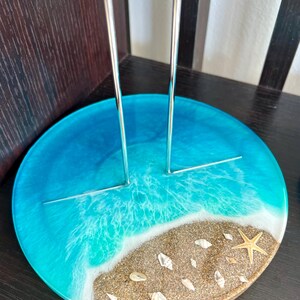 May include: A round, blue and brown resin base with a silver metal stand. The base features a beach scene with white waves, brown sand, and white seashells. A gold starfish is on the sand.