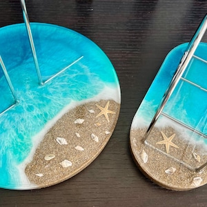May include: Two resin coasters with a beach theme. The coasters are round and have a blue and white ocean wave design with a sandy beach and seashells. One coaster has a metal stand for holding napkins or paper towels.