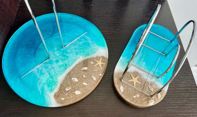 May include: Two resin coasters with a beach theme. The coasters are round and have a blue and white ocean wave design with a sandy beach and seashells. One coaster has a metal stand for holding napkins or paper towels.