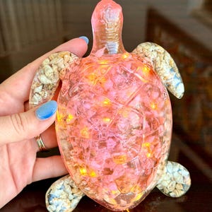 May include: A translucent resin sea turtle figurine featuring a pink-tinted shell filled with small pink stones and embedded warm yellow LED lights. The turtle's flippers are decorated with a collection of small, natural-colored seashells.