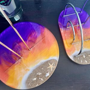 May include: Two round resin coasters with a sunset design. The coasters are purple, orange, and yellow with white sand and seashells. One coaster has a metal holder for incense sticks.